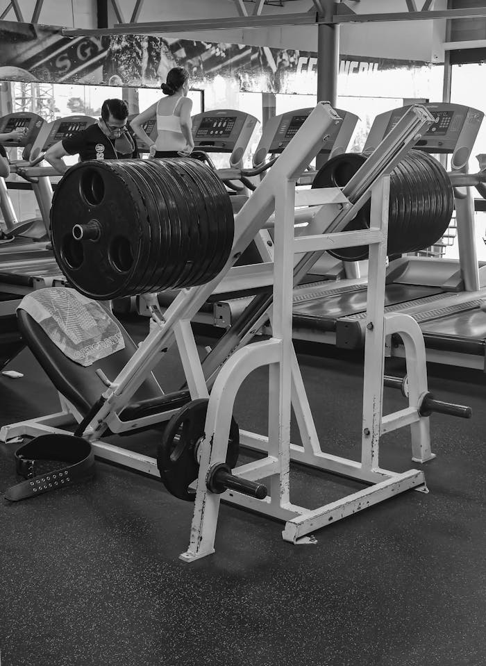 heros-img Black and white photo of a leg press machine in a gym with treadmills and people exercising.